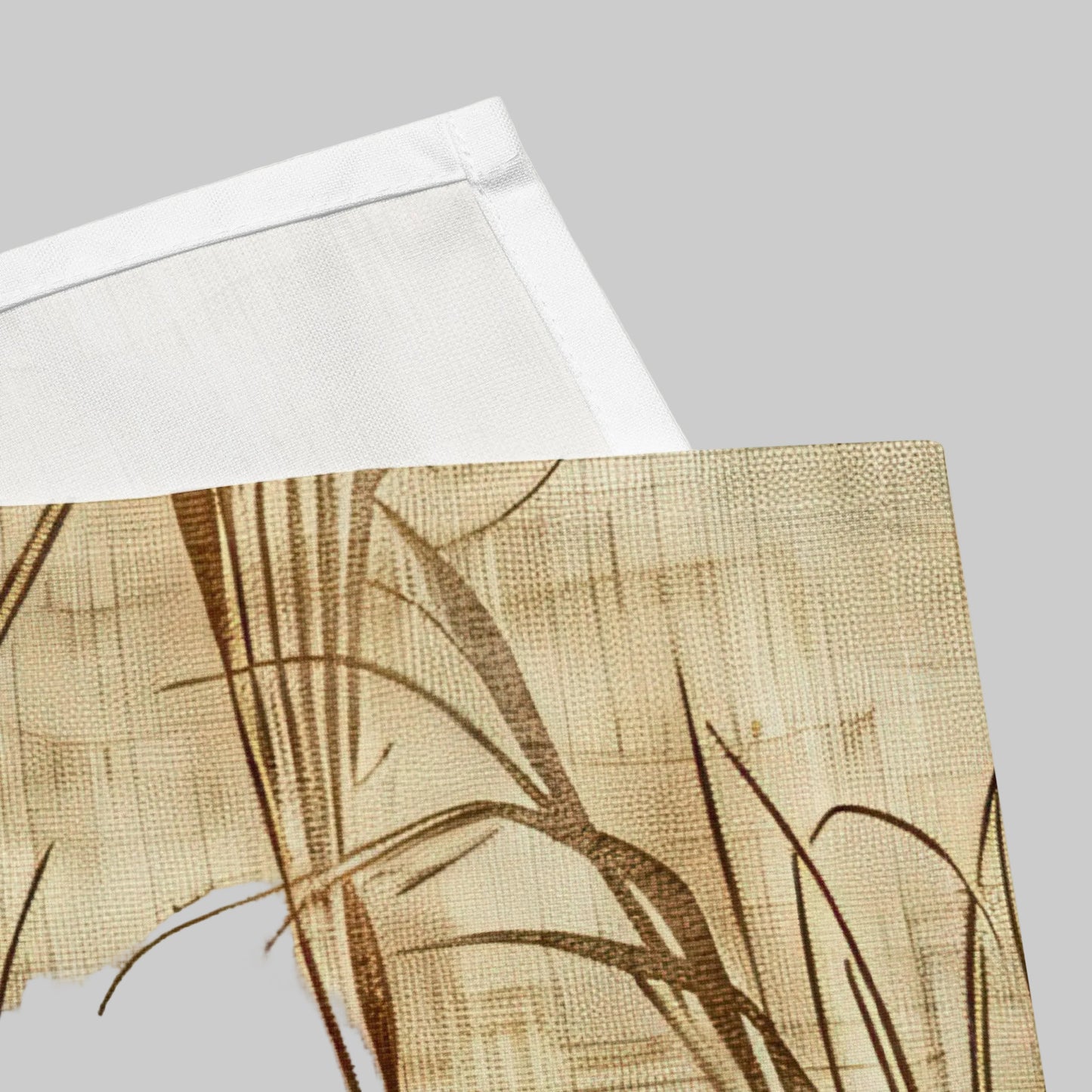A close-up of a beige woven Napkin with a brown reed or grass pattern, partially folded over to show a white underside, on a white background.