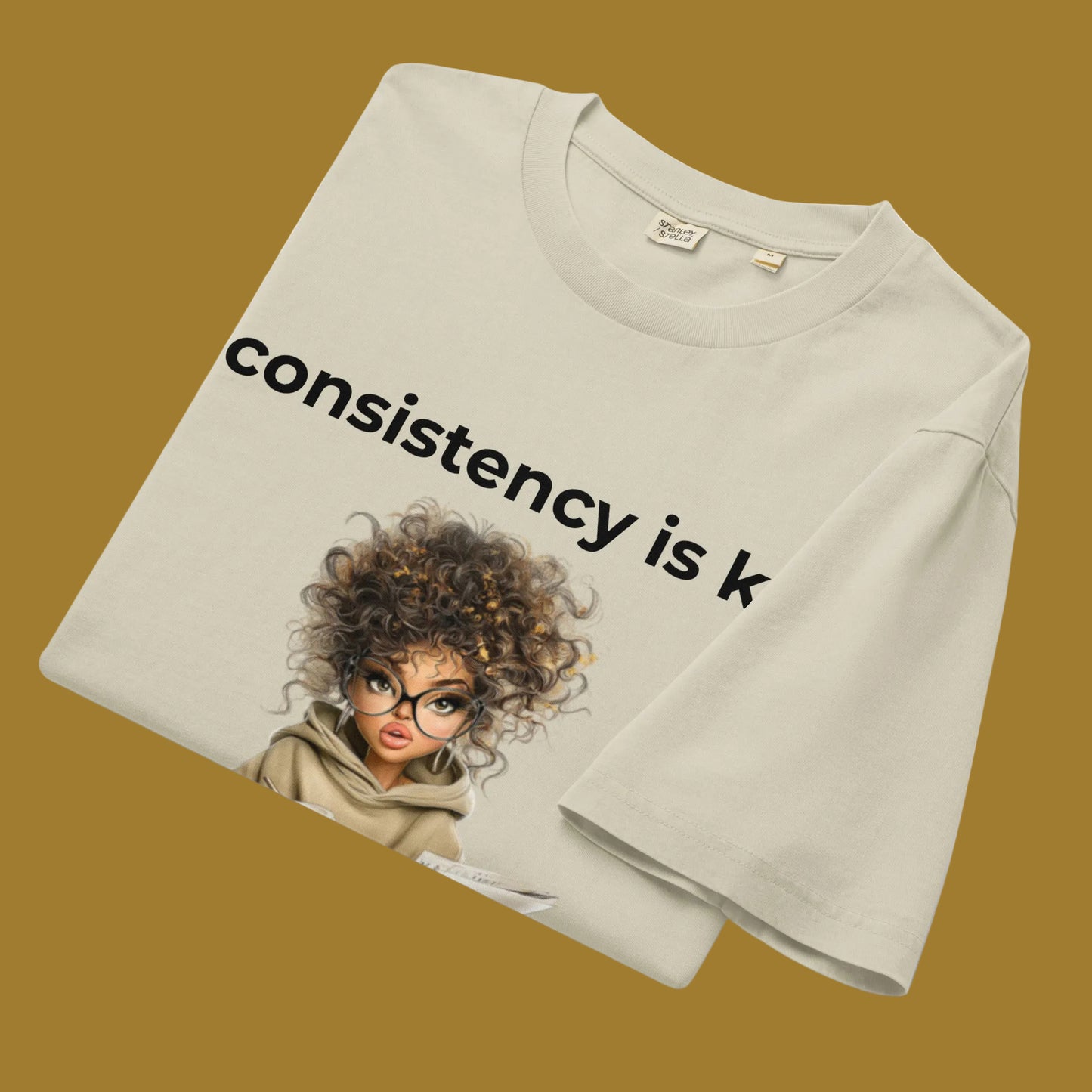 Beige t-shirt with a graphic of a woman with text 'consistency is k' on a goldbackground